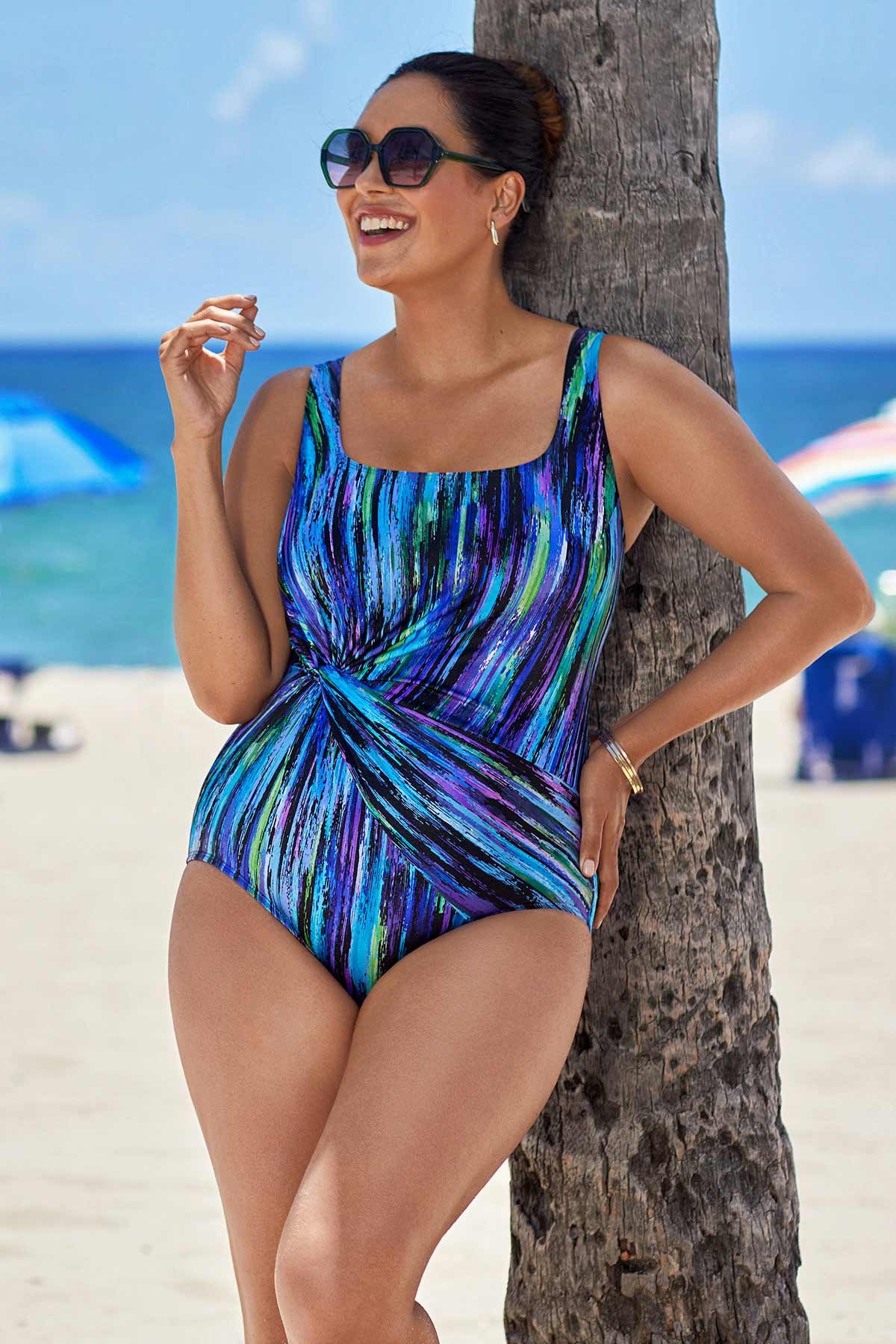 Woman on beach wearing a multicolored streak printed one-piece swimsuit with a side twist